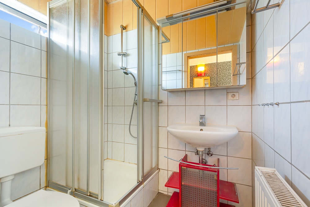 Kleines Badezimmer mit Dusche, Toilette, Waschbecken, Spiegel und Heizung.