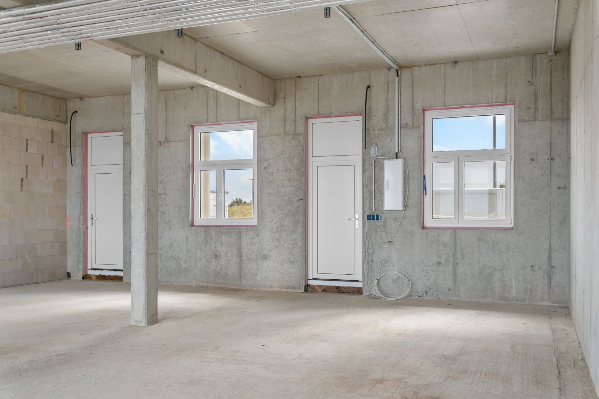 Leerer Raum mit Betonwänden, zwei Türen, zwei Fenstern und sichtbaren Leitungen.