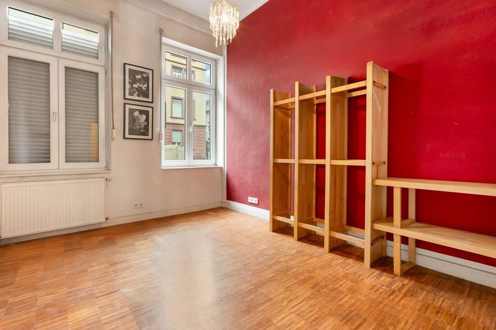 Leerer Raum mit roter Wand, Holzboden, Regal, zwei Fenstern und Kronleuchter.