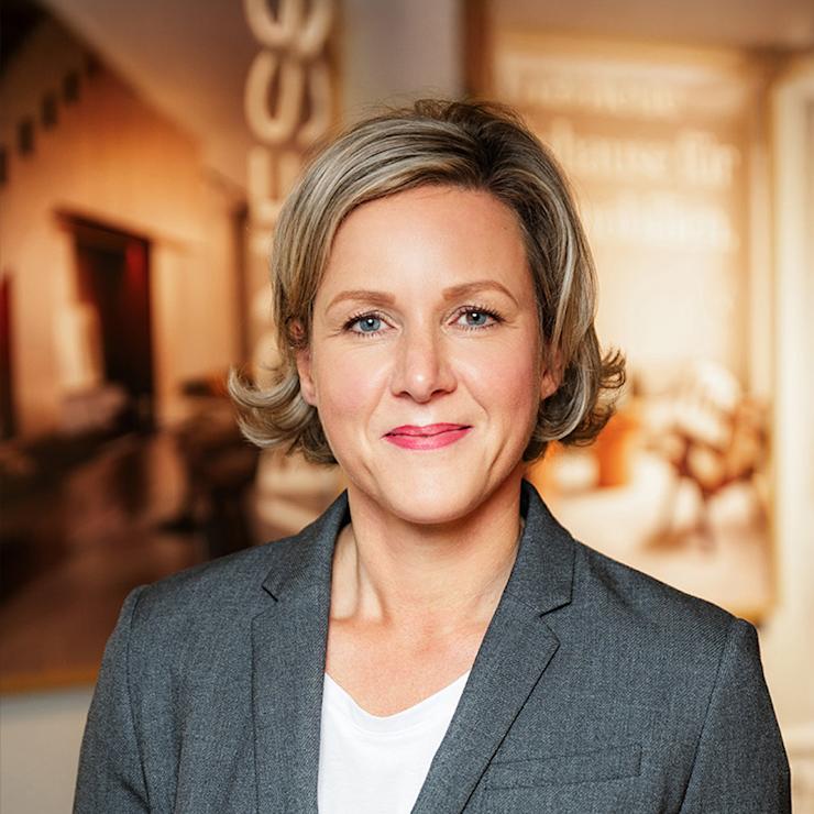 Frau mit grauem Blazer steht in einem hellen Innenraum.