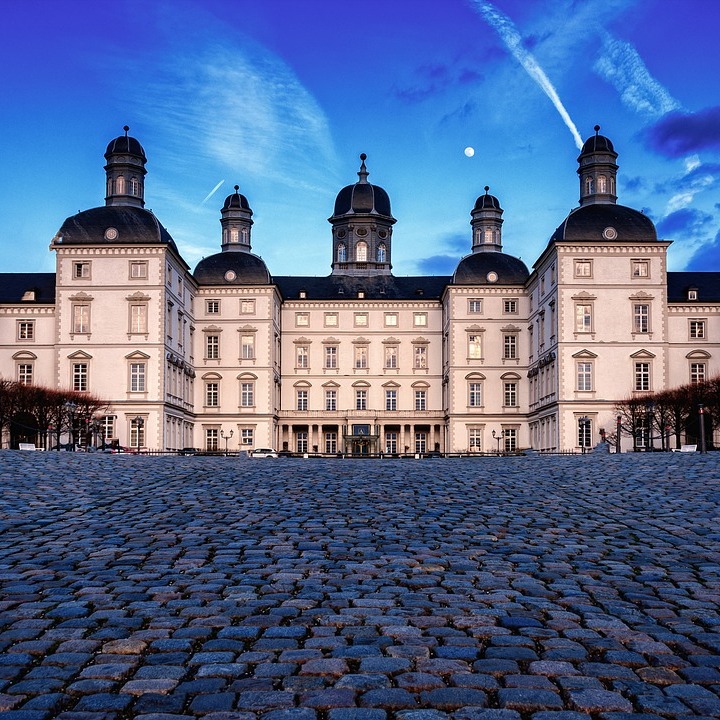 Großes, helles Gebäude mit vier Türmen, Kopfsteinpflaster im Vordergrund, blauer Himmel.