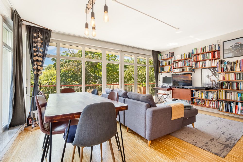 Helles Wohnzimmer mit großem Fenster, Esstisch, grauem Sofa und Bücherregalen.