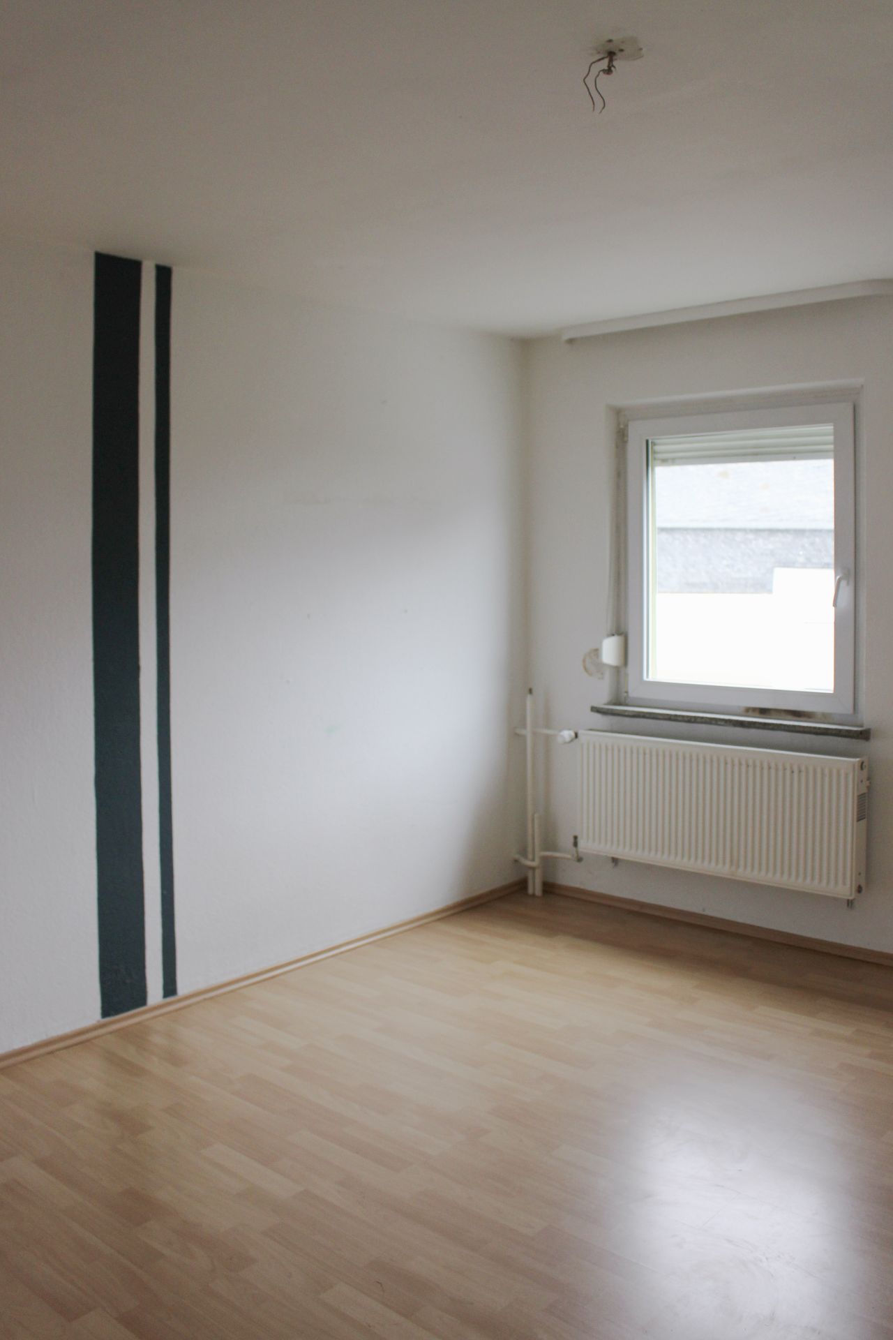 Leeres Zimmer mit hellen Holzboden, weißer Wand, Fenster und Heizkörper.