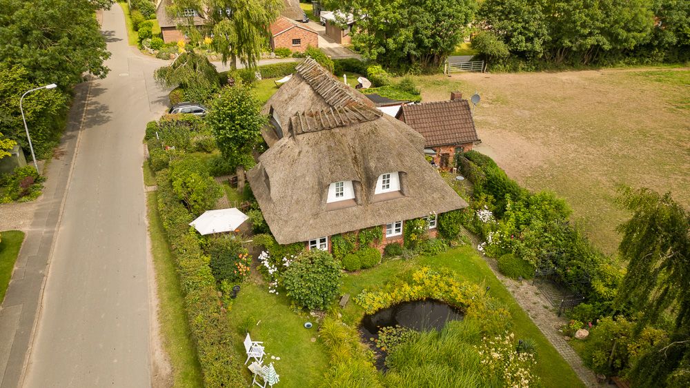 Reetgedecktes Haus mit Garten, Teich und Terrasse an einer ruhigen Straße, umgeben von Grün.