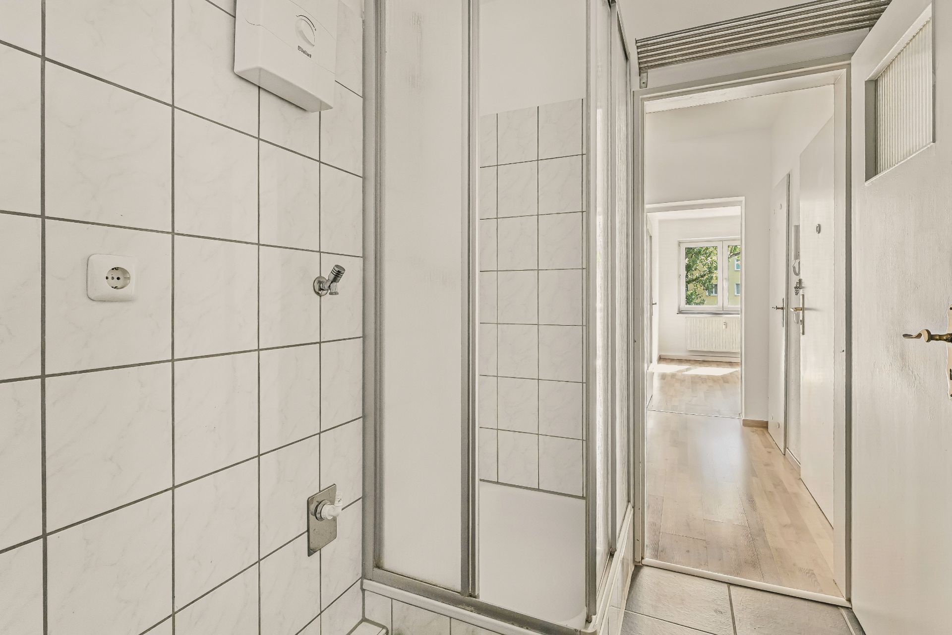 Badezimmer mit gefliester Dusche, offenem Türrahmen und Blick in einen hellen Flur mit Holzfußboden.