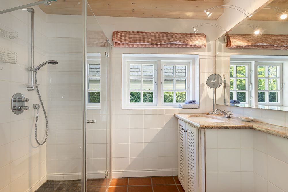 Helles Badezimmer mit Dusche, Waschbecken, großem Spiegel und zwei Fenstern.