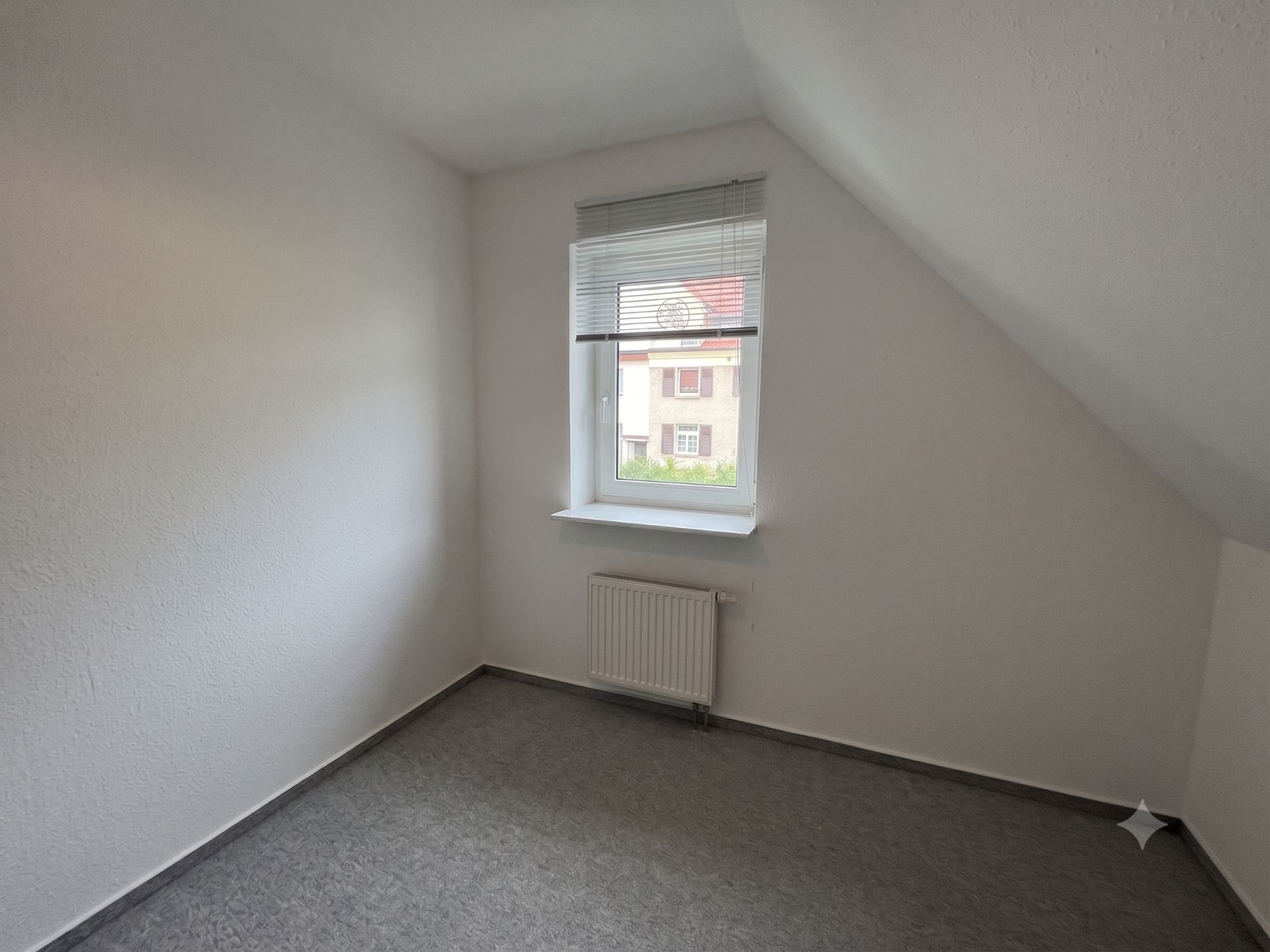 Kleines, leeres Zimmer mit Fenster, Jalousie und Heizkörper, grauer Teppichboden.