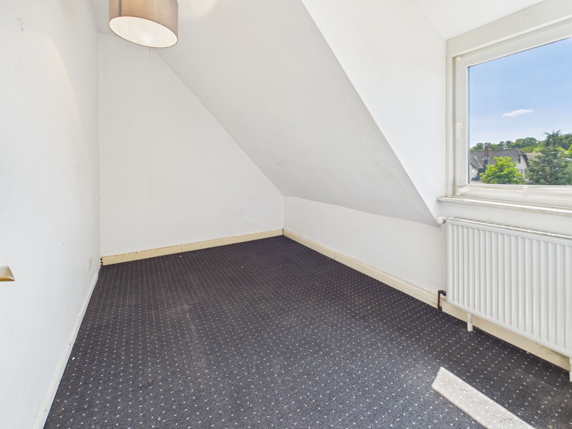 Kleines Zimmer mit Dachschräge, Teppichboden, Fenster und Heizkörper.
