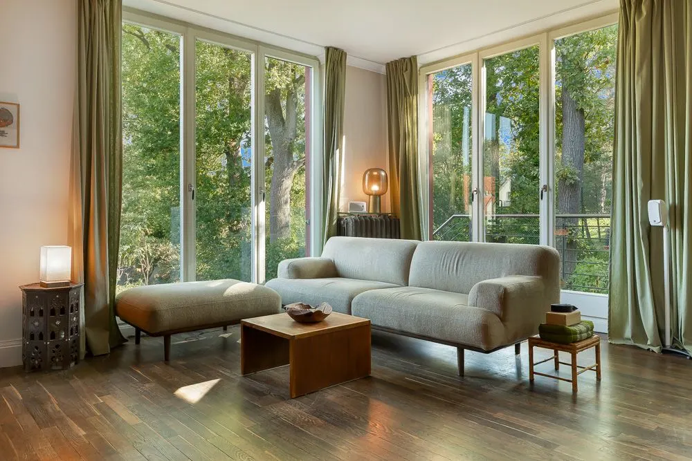Helles Wohnzimmer mit Sofa, Hocker, Couchtisch und großen Fenstern mit Blick auf Bäume.