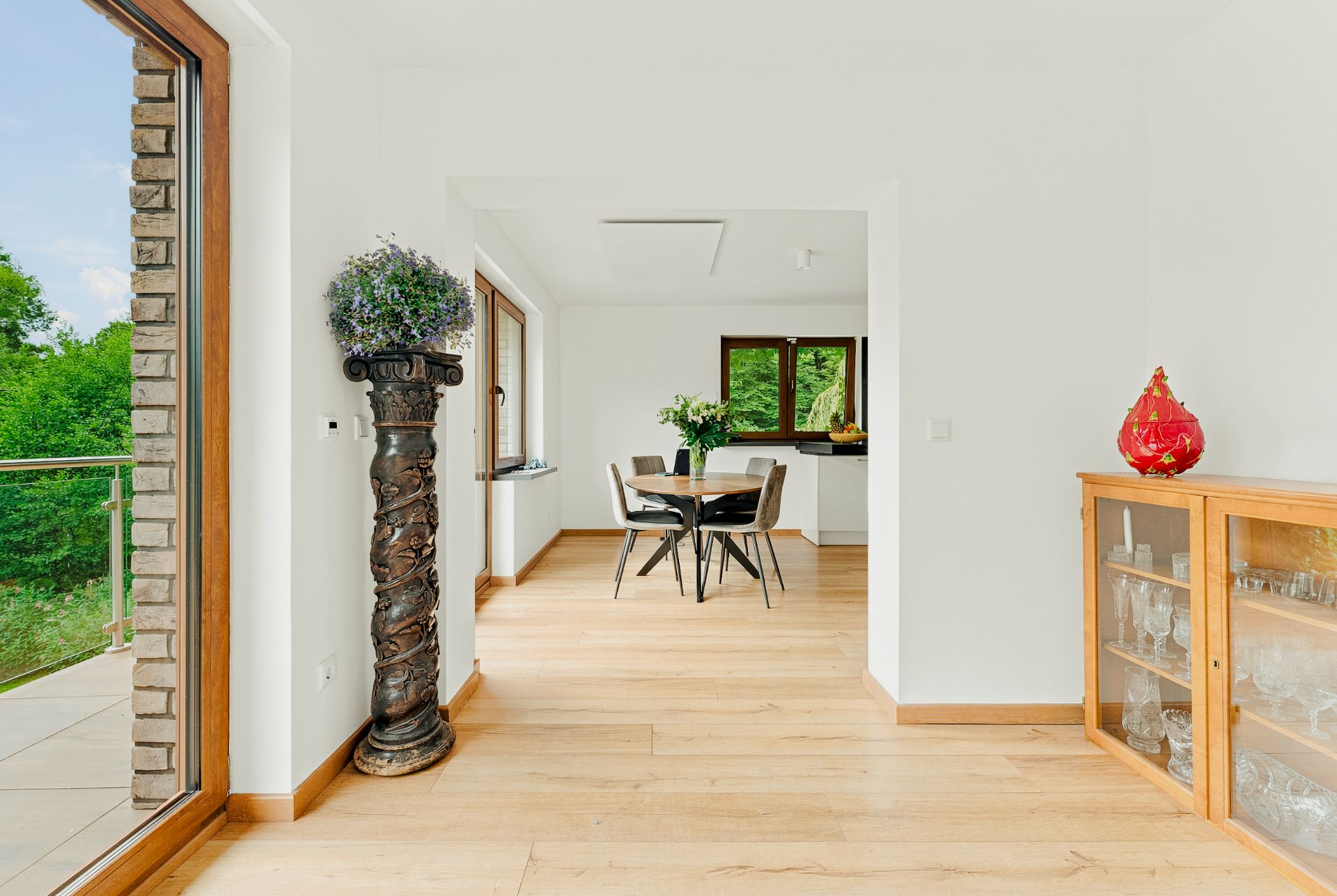 Heller Raum mit Holzboden, Esstisch, Glasmöbel und großen Fenstern mit Blick ins Grüne.