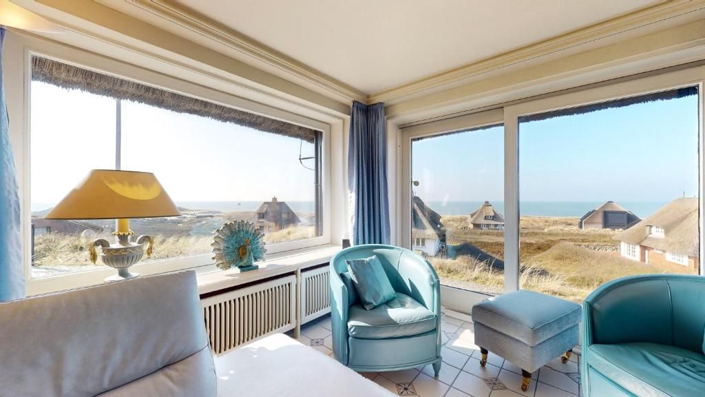 Wohnzimmer mit großen Fenstern, zwei Sesseln, Meerblick und Häusern mit Reetdach im Hintergrund.