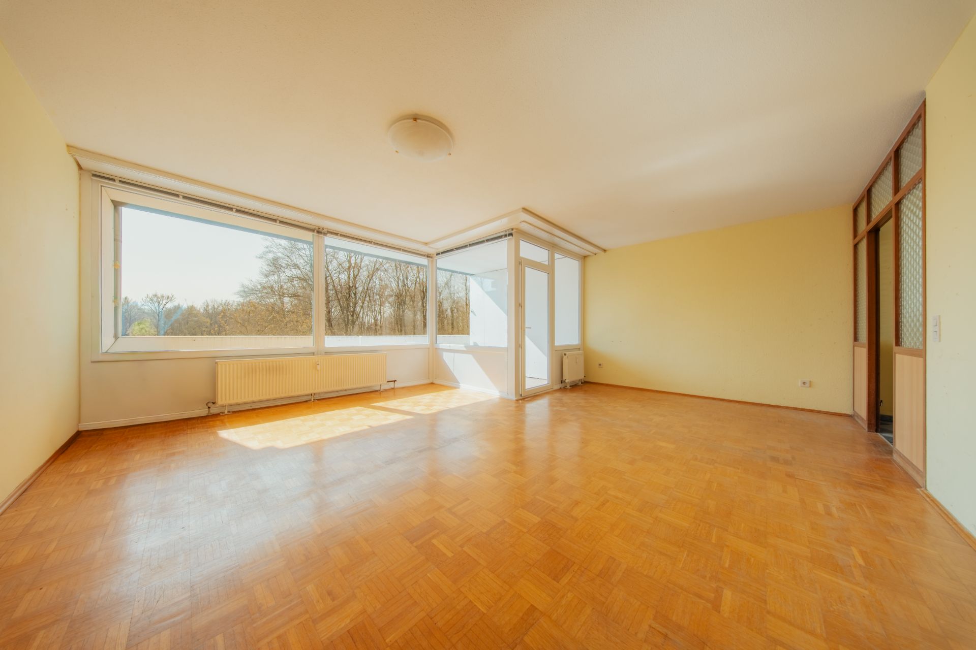 Helles, leeres Wohnzimmer mit großen Fenstern und Parkettboden.
