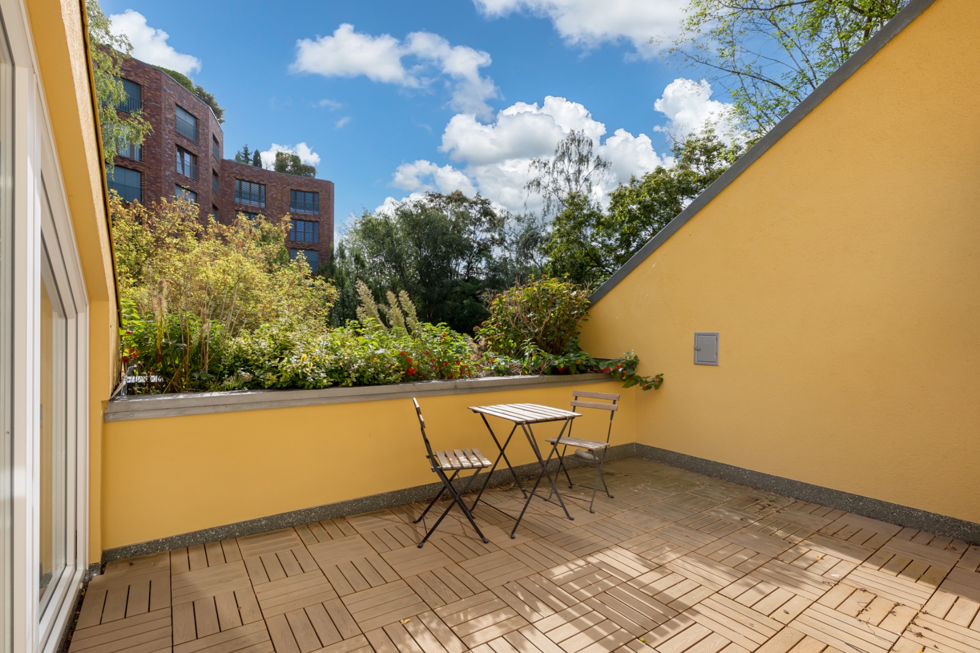 Helle Terrasse mit zwei Stühlen, kleinem Tisch und Pflanzen an einer gelben Wand.