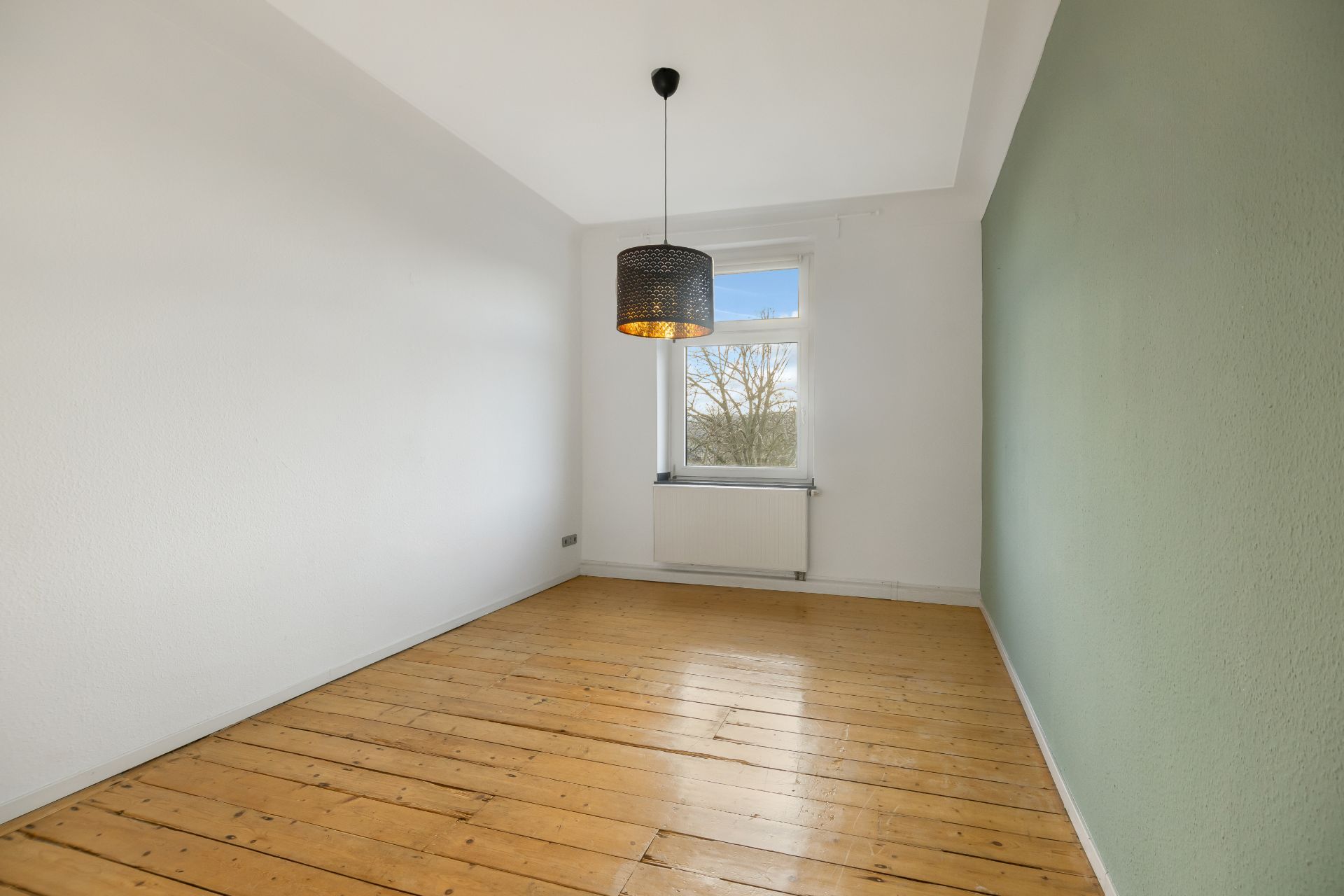 Leerer Raum mit Holzboden, weiß-grünen Wänden, Fenster und schwarzer Deckenlampe.