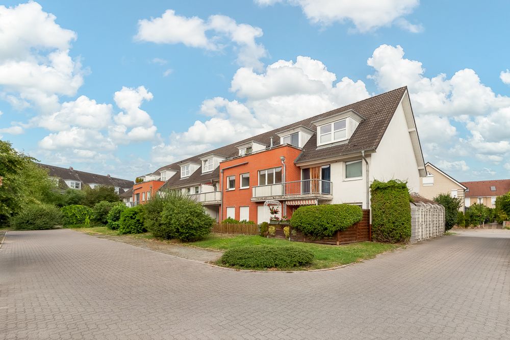 Mehrfamilienhaus mit roter Fassade, Balkon und Vorgarten an einer gepflasterten Straße.