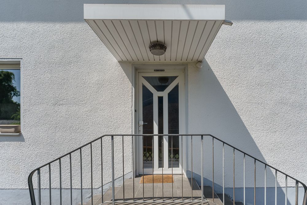 Weiße Haustür mit Glas, Überdachung und Metallgeländer vor einem Wohnhaus.