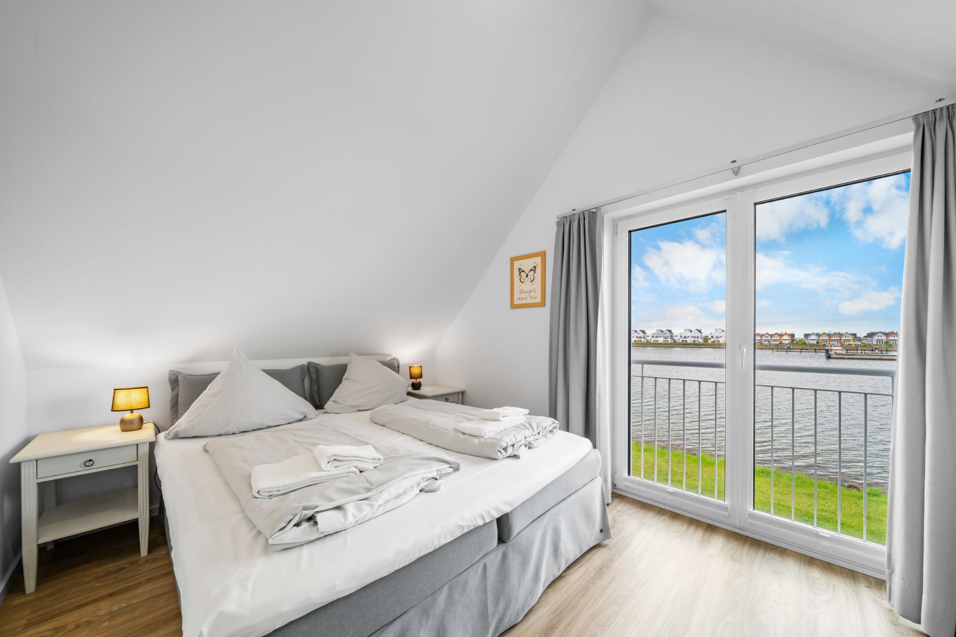 Helles Schlafzimmer mit Doppelbett, zwei Nachttischen und großen Fenstern mit Blick auf Wasser und Häuser.