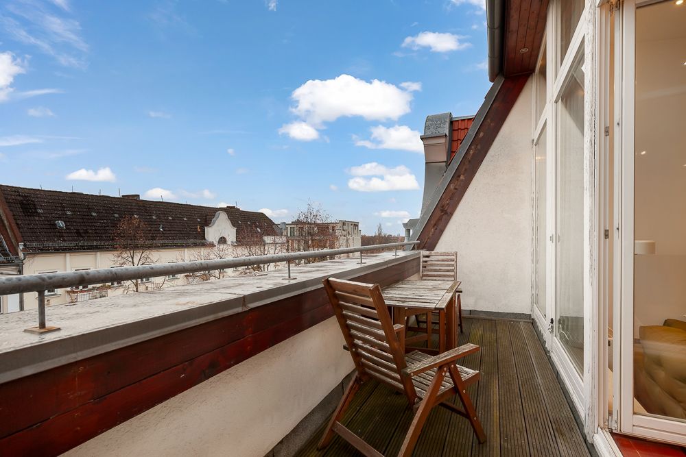 Balkon mit Holztisch und zwei Stühlen, Blick auf Nachbarhäuser und blauen Himmel.