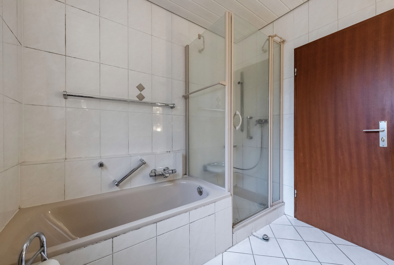 Badezimmer mit Badewanne, Dusche mit Glastür und brauner Holztür.