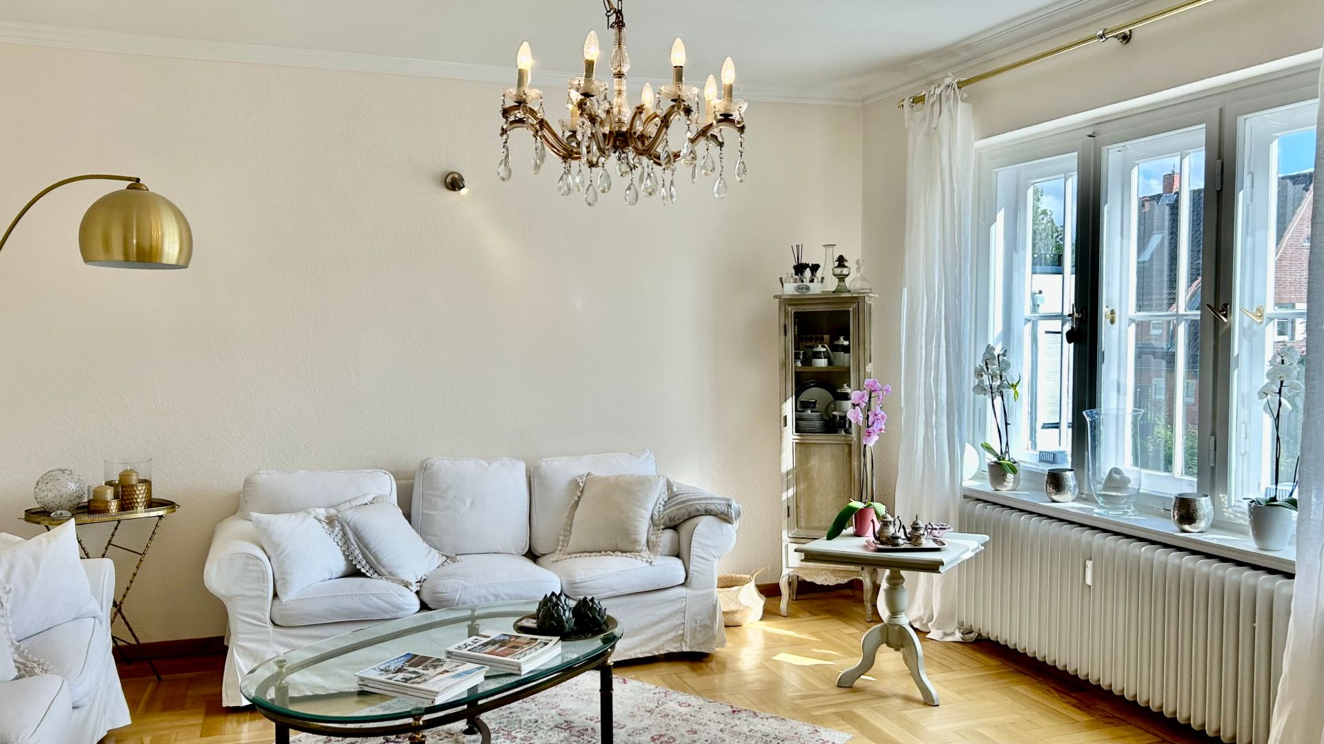 Helles Wohnzimmer mit weißen Sofas, Glas-Couchtisch, Kronleuchter und großen Fenstern.