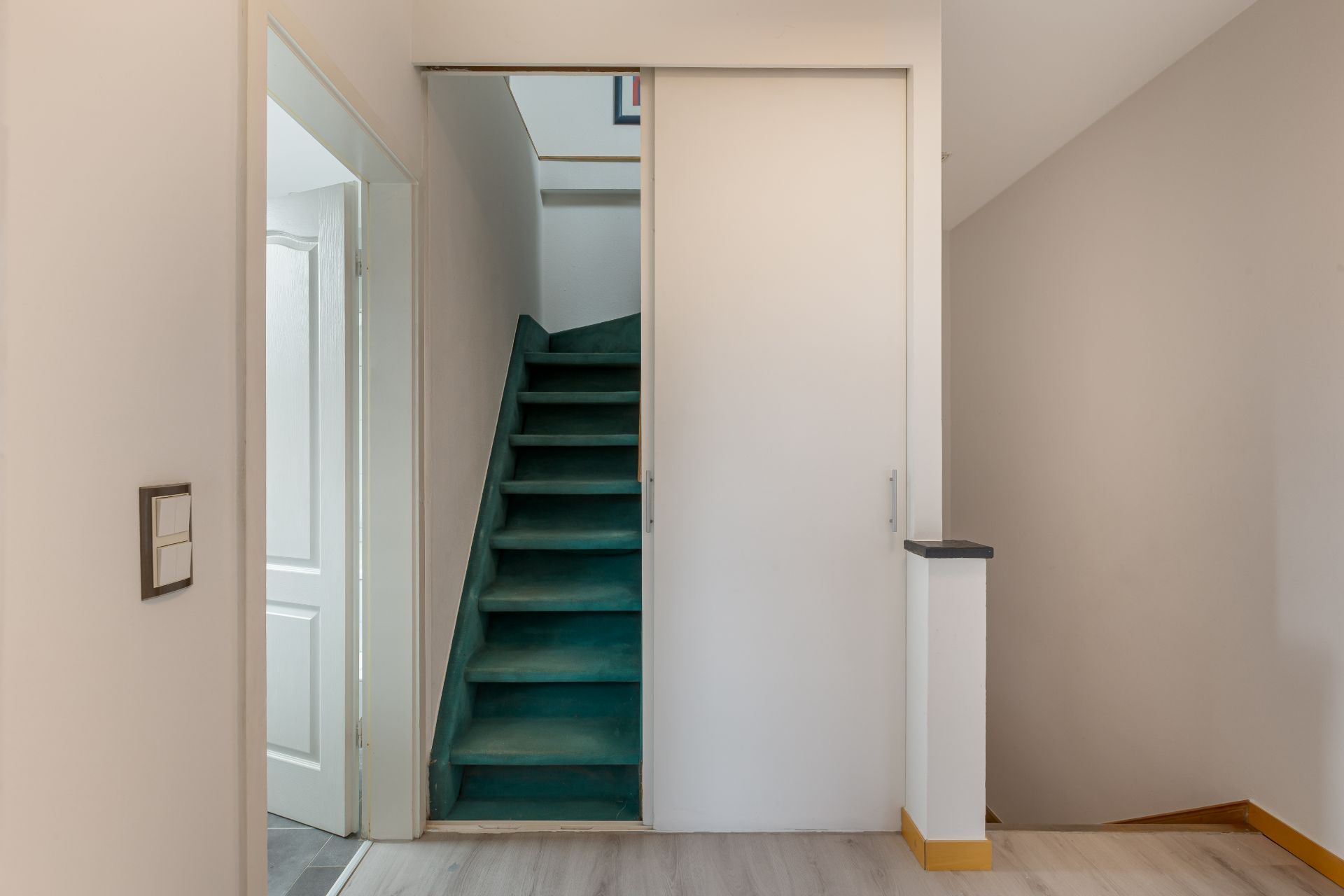Flur mit grüner Treppe hinter einer Schiebetür und hellen Wänden.