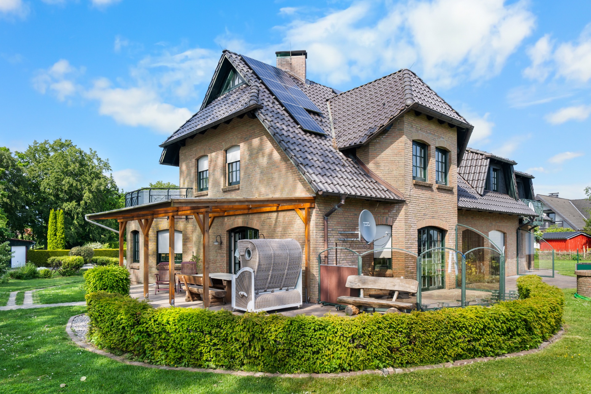 Großes Einfamilienhaus mit Terrasse, Sitzmöbeln, Solarzellen und Garten bei Sonnenschein.