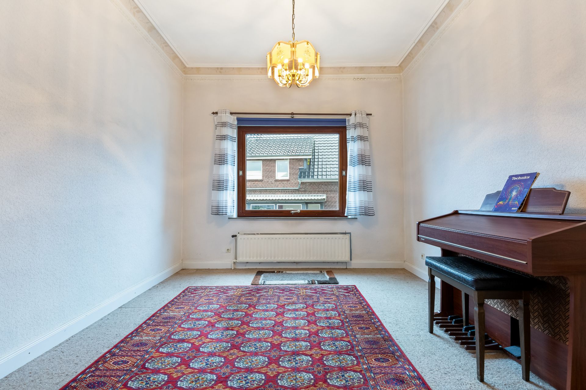 Kleines Zimmer mit rotem Teppich, Fenster, Vorhängen, Hängelampe und einem Klavier rechts.
