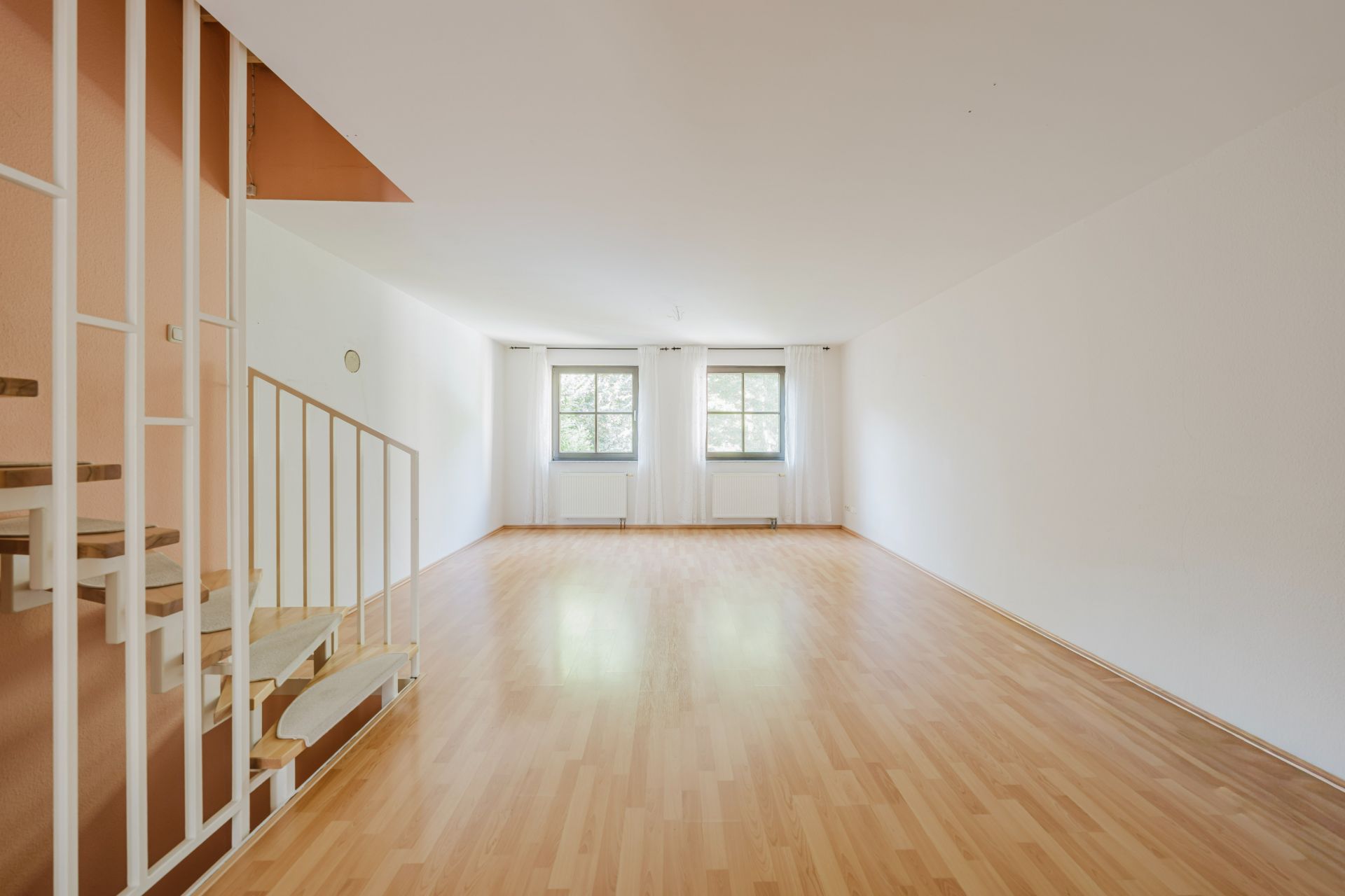 Leeres Wohnzimmer mit Holzfußboden, zwei Fenstern und einer Treppe links.