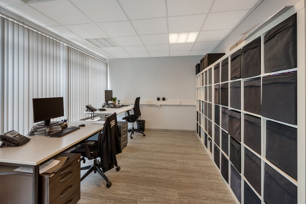 Büro mit zwei Schreibtischen, Computern, Stühlen und Regalwand mit schwarzen Boxen.