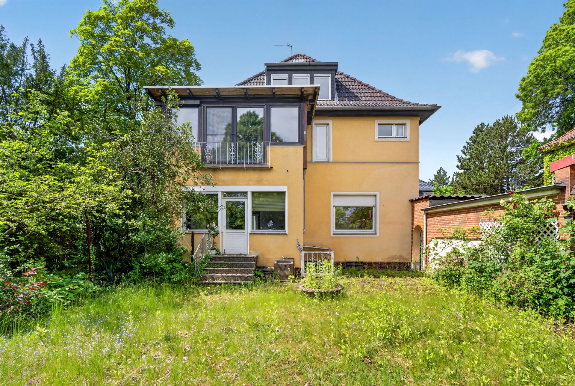 Gelbes Wohnhaus mit Wintergarten und Garten im Vordergrund, umgeben von Bäumen.