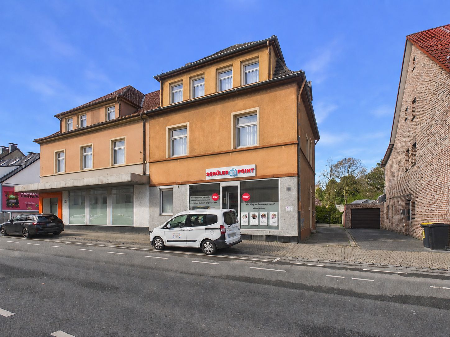 Zweistöckiges Wohnhaus mit Laden im Erdgeschoss, parkende Autos davor, Straße und Gehweg sichtbar.