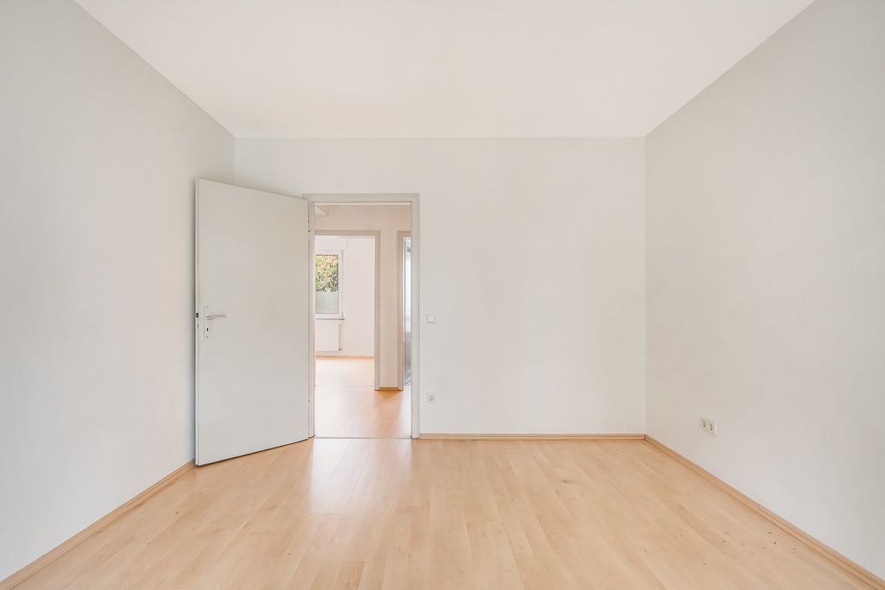 Leeres Zimmer mit hellem Holzboden, offener Tür und Blick in einen weiteren Raum mit Fenster.
