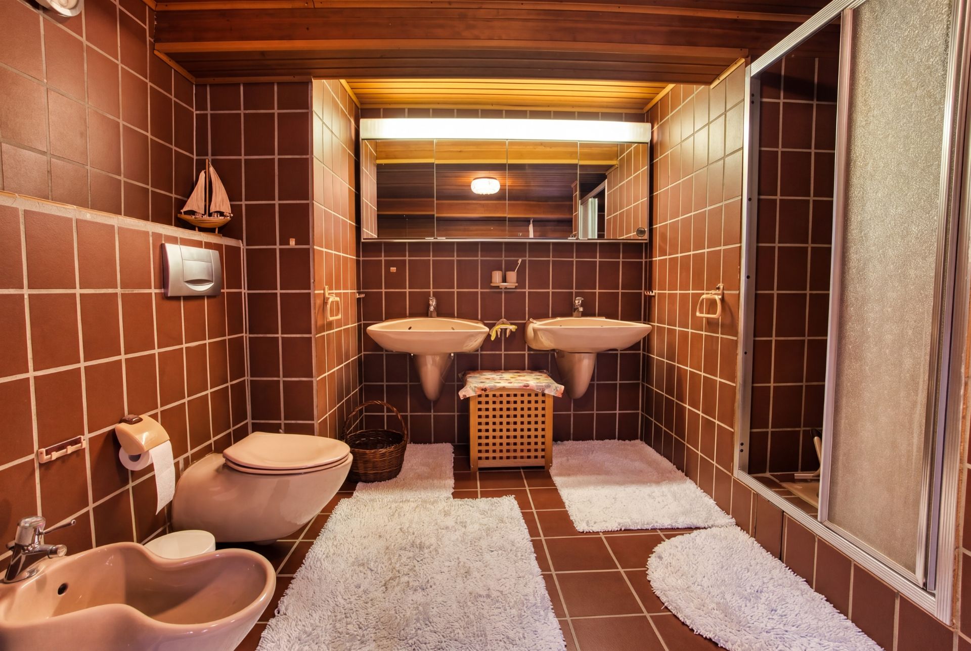Badezimmer mit braunen Fliesen, zwei Waschbecken, Dusche, Toilette, Bidet und weißen Teppichen.