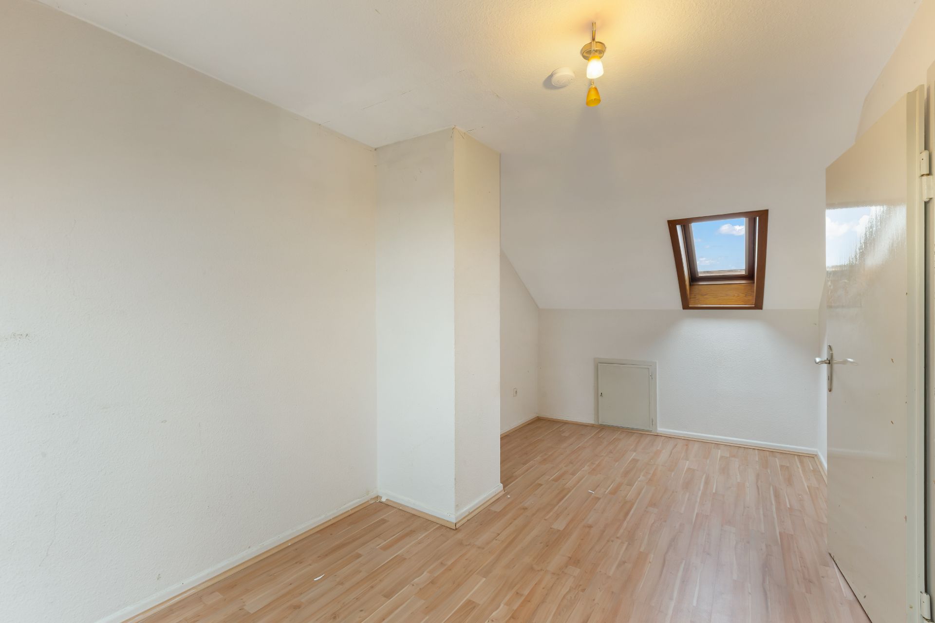 Leeres Zimmer mit Holzfußboden, Dachschräge und kleinem Fenster im Dach.