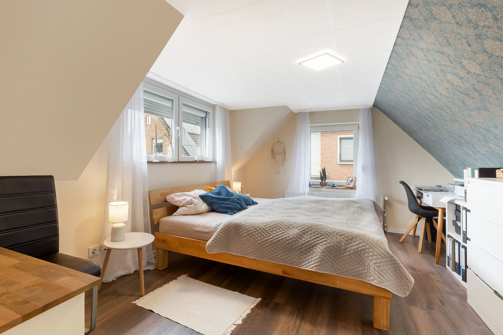 Helles Schlafzimmer mit Doppelbett, Schreibtisch, Sesseln und zwei Fenstern auf Dachgeschoss-Ebene.