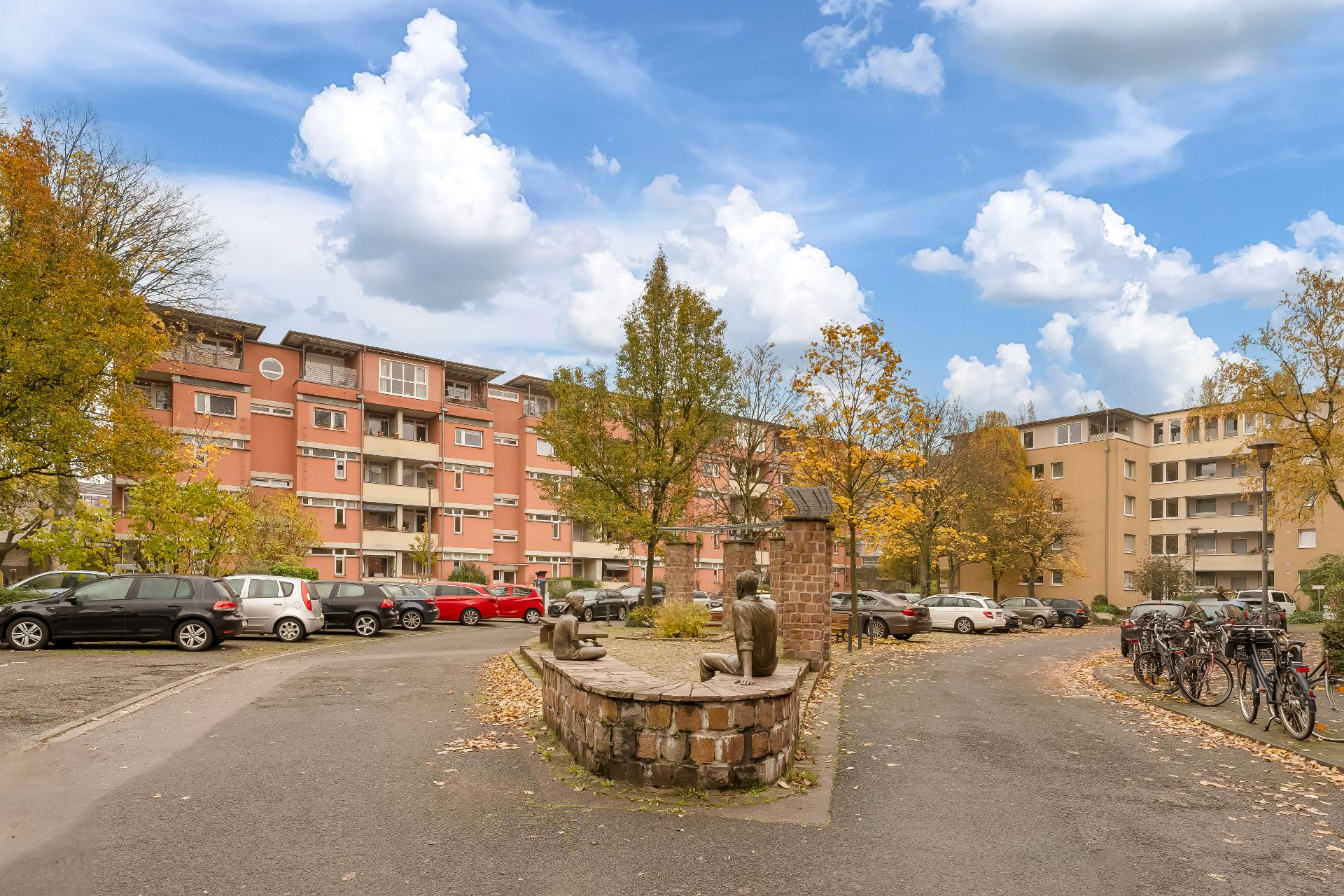 Wohngebäude mit geparkten Autos, Fahrrädern und einer Steinskulptur in einer herbstlichen Umgebung.