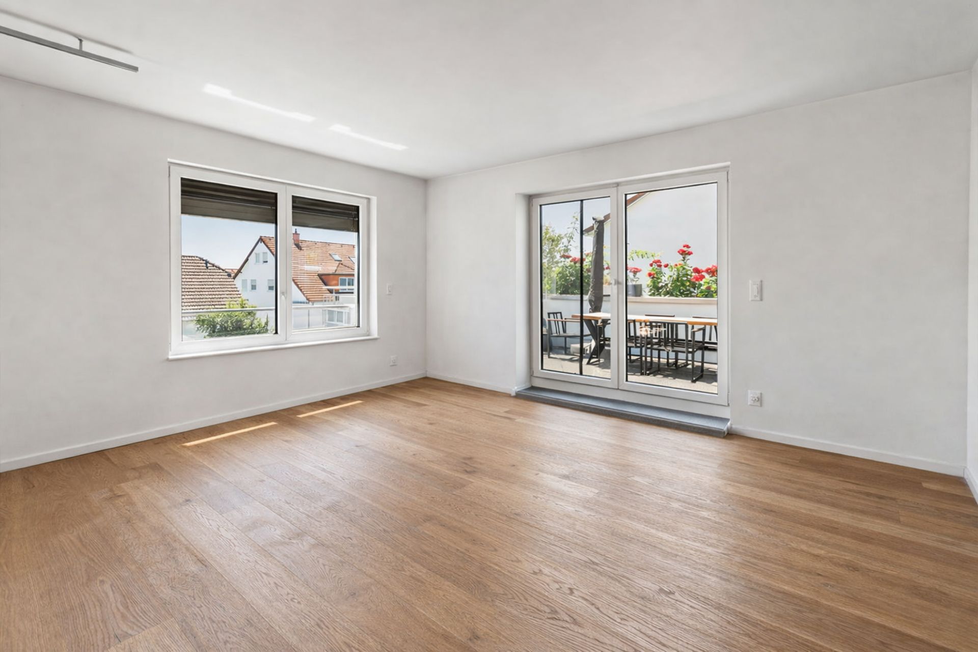 Helles, leeres Zimmer mit Holzboden, zwei Fenstern und Zugang zu einem Balkon mit Tisch und Stühlen.