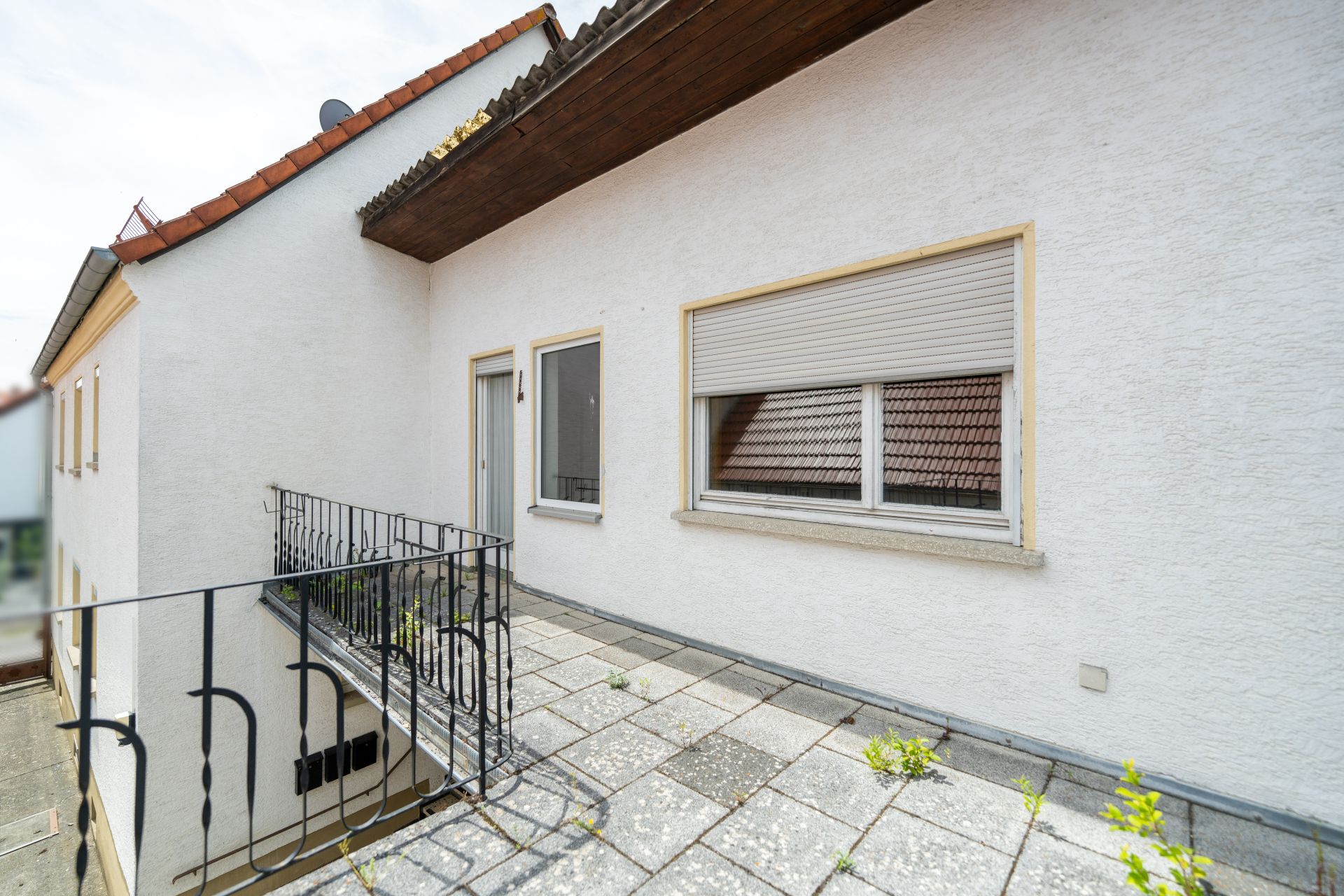 Balkon mit Fliesenboden, Metallgeländer und zwei Fenstern an einem weißen Wohnhaus.