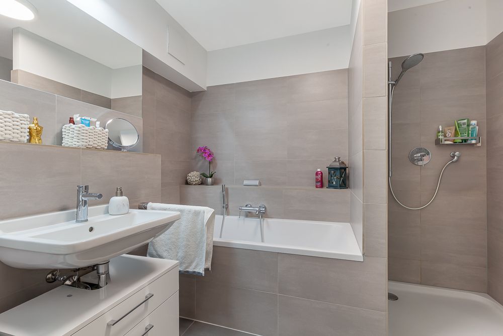 Modernes Badezimmer mit Waschbecken, Badewanne und Dusche, graue Fliesen und einigen Dekorationen.