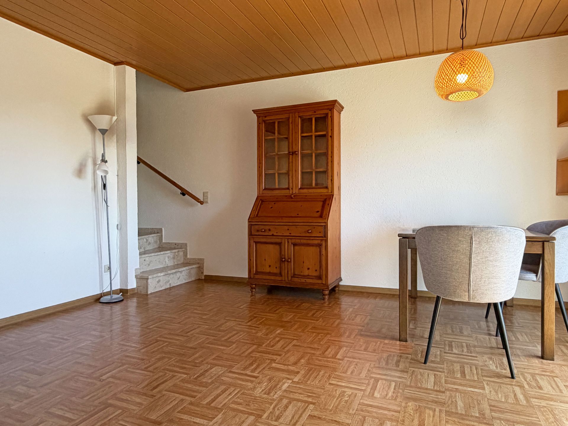 Wohnzimmer mit Holzschrank, Esstisch, zwei Stühlen, Stehlampe und Treppe.