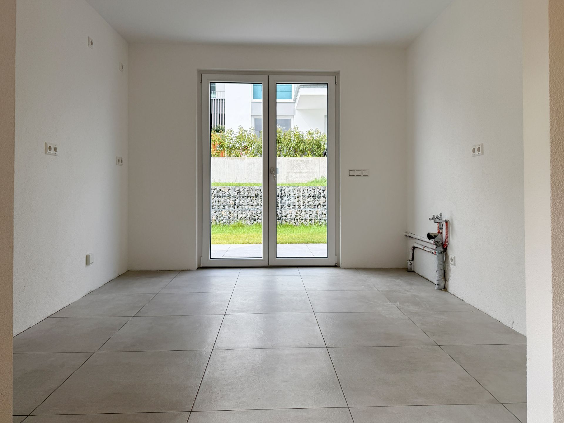 Leerer Raum mit Fliesenboden und großer Glastür zum Garten.