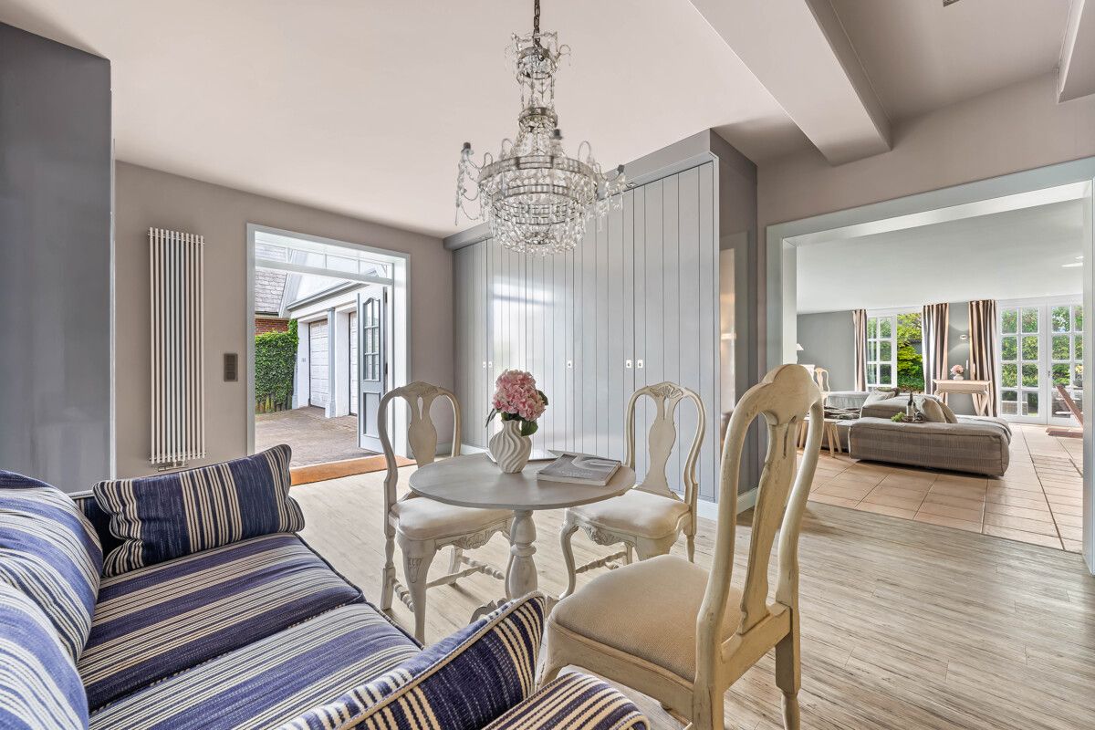 Heller Essbereich mit rundem Tisch, Stühlen, Sofa und Kronleuchter, daneben offenes Wohnzimmer.