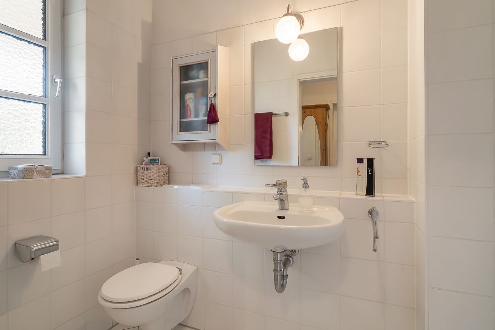 Helles Bad mit WC, Waschbecken, Spiegel, Fenster und Hängeschrank an weiß gefliester Wand.