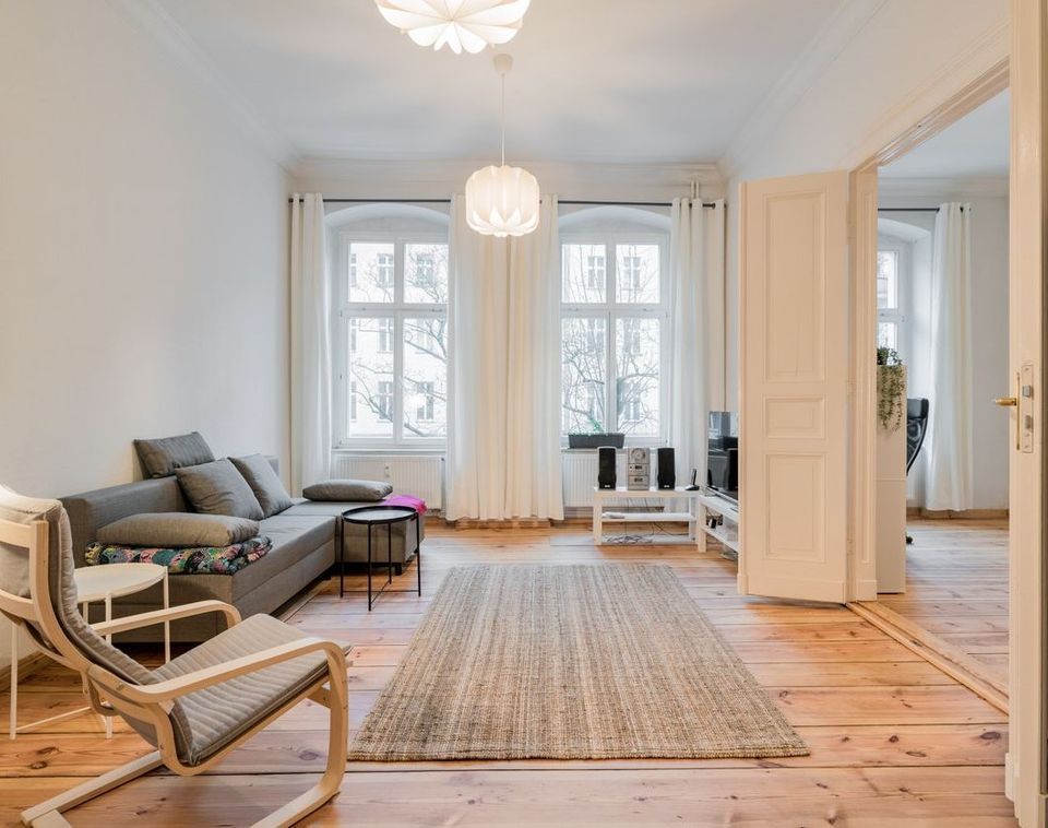 Helles Wohnzimmer mit Sofa, Sessel, Teppich und großen Fenstern mit weißen Vorhängen.