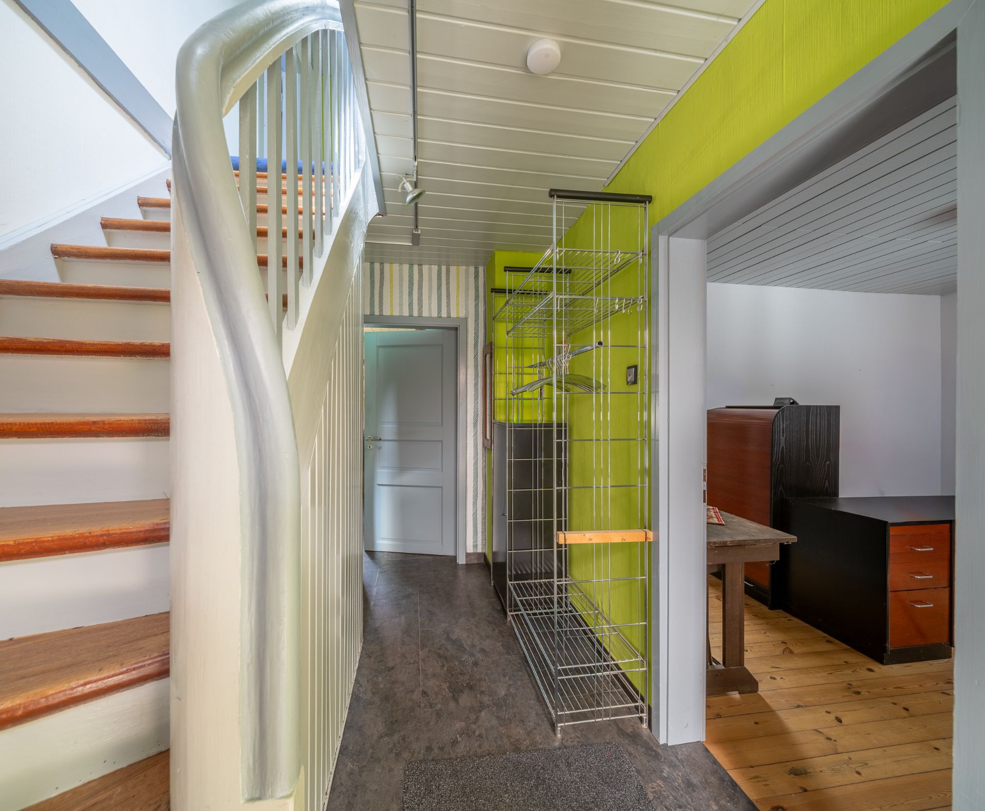 Flur mit Treppe, Garderobe und Blick in ein angrenzendes Zimmer mit Schreibtisch.