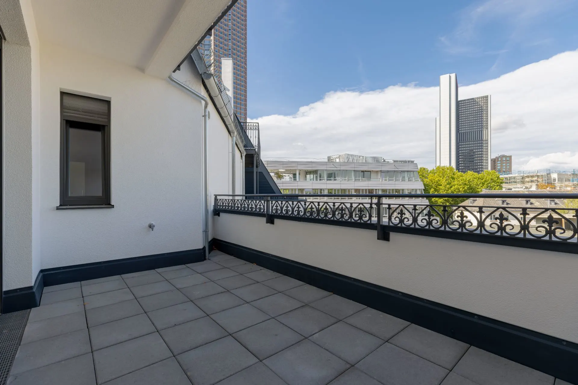 Großer Balkon mit Fliesenboden und Metallgeländer, Blick auf moderne Gebäude.