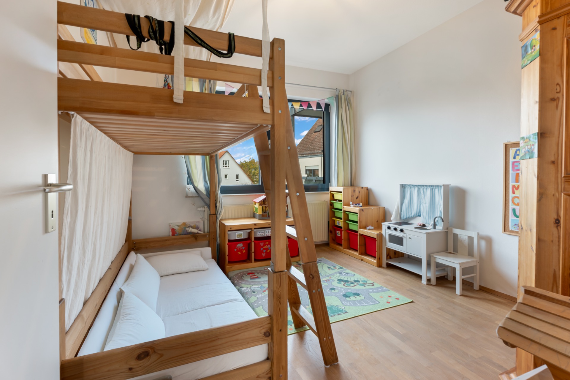 Helles Kinderzimmer mit Hochbett, Spielzeugregalen und Spielküche auf Holzboden.