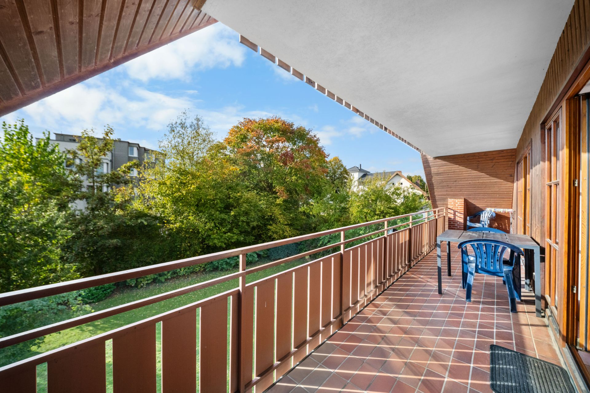 Großer Balkon mit Fliesenboden, Tisch und Stühlen, Blick auf Bäume und Nachbarhäuser.