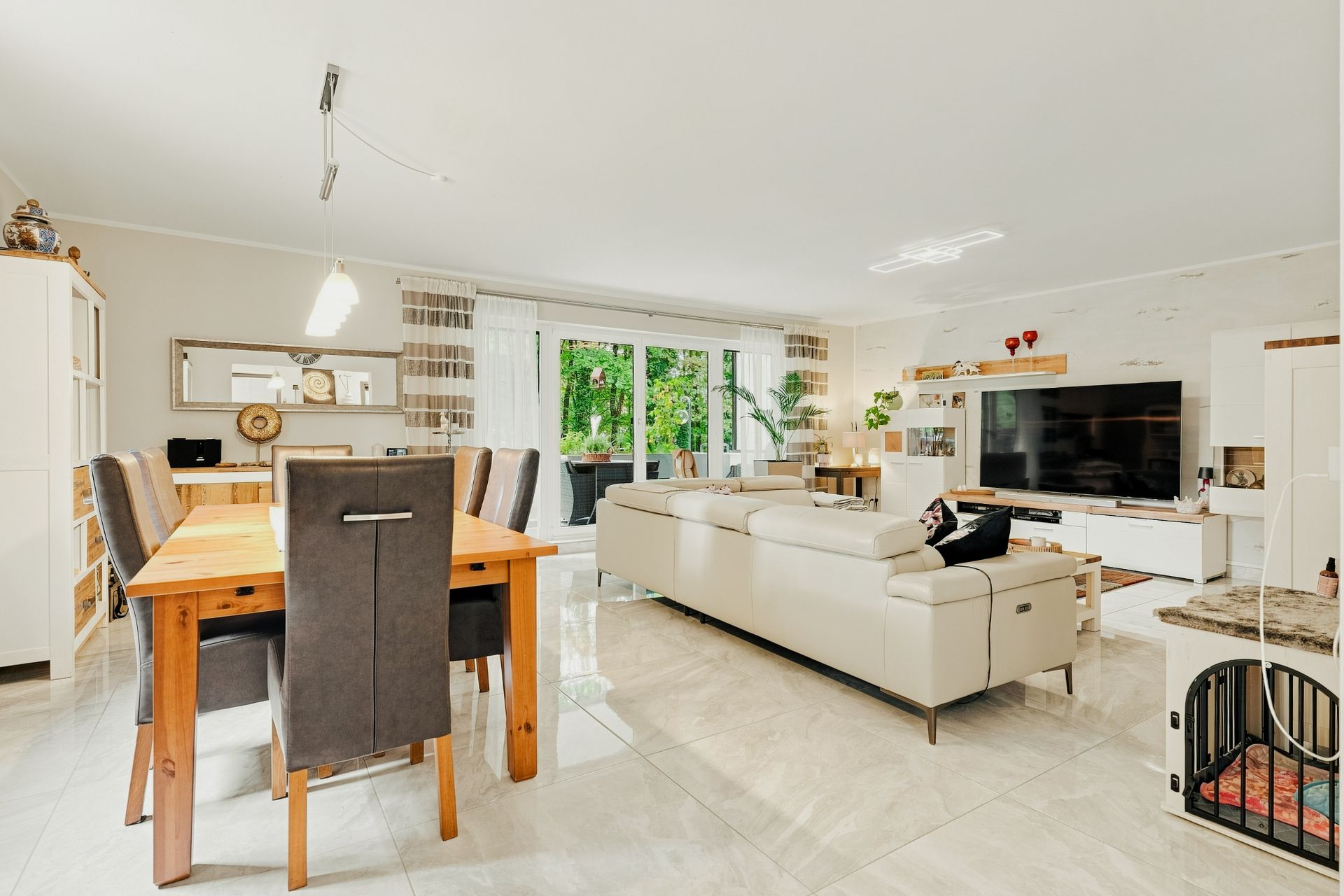 Helles Wohnzimmer mit Essbereich, großem Sofa, Esstisch, Fernseher und Blick auf eine Terrasse mit Garten.