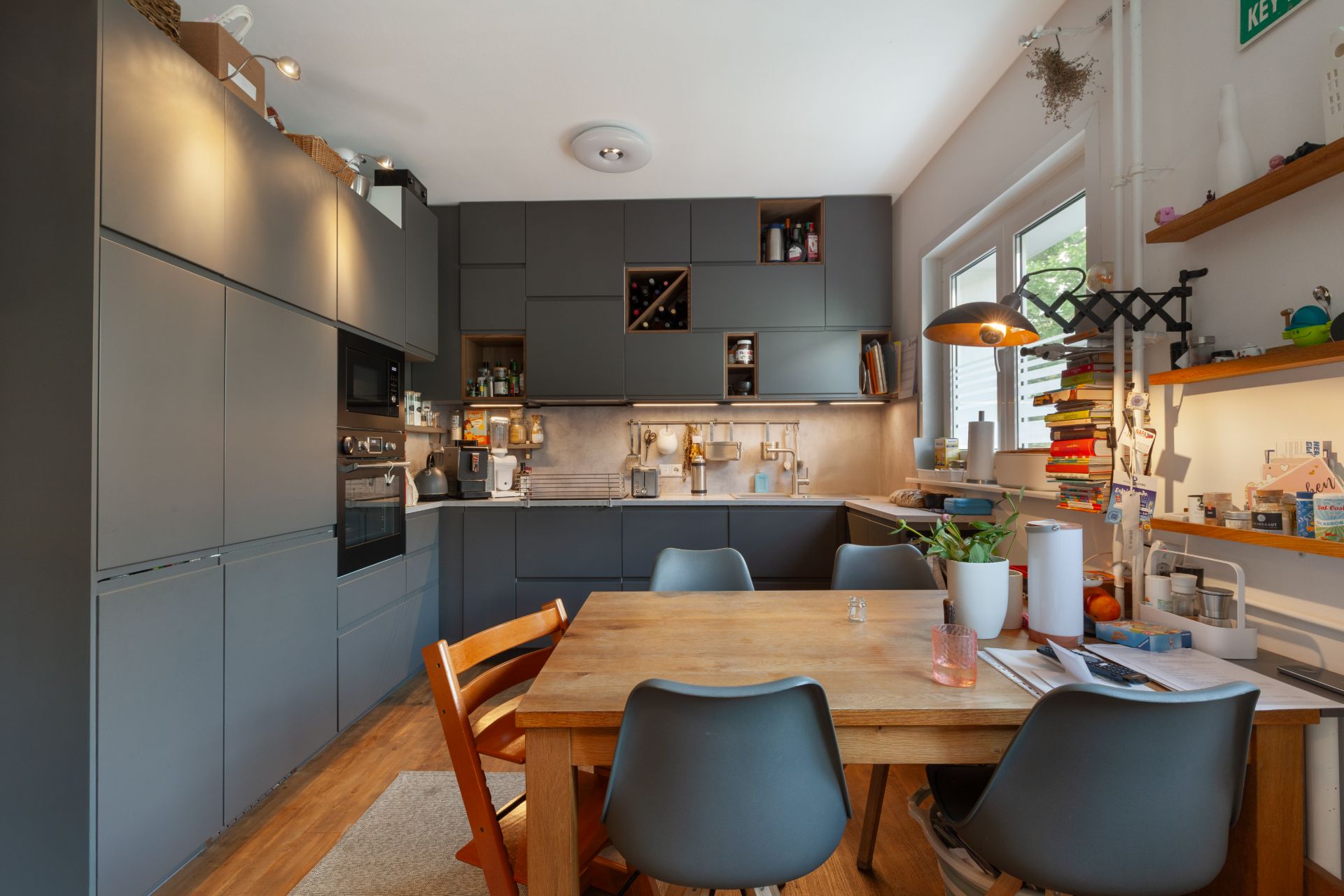 Moderne Küche mit grauen Schränken, Holztisch und vier Stühlen, Fenster rechts, Regale und Lampen an der Wand.